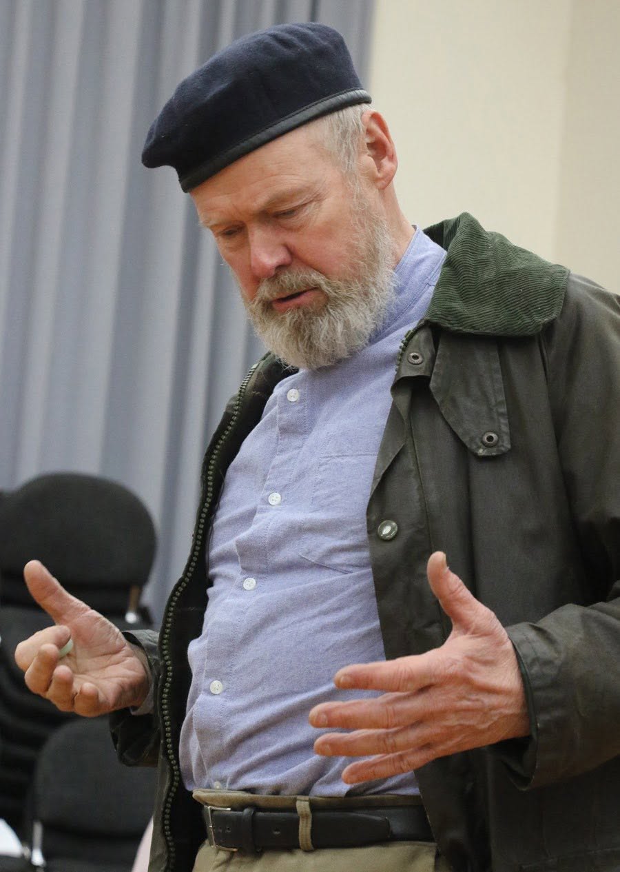 An As You Like It performer rehearsing in a dark cap and green jacket